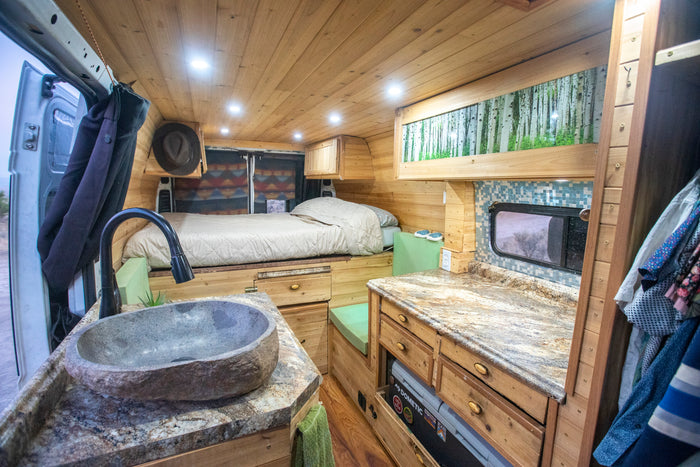 Cozy RV rental interior with custom wood, a bed, stone sink, and granite counter kitchen. Perfect camper van experience.