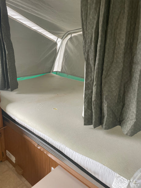Pop-up RV rental bunk interior, with a mattress, gray curtains, and white fabric walls visible.