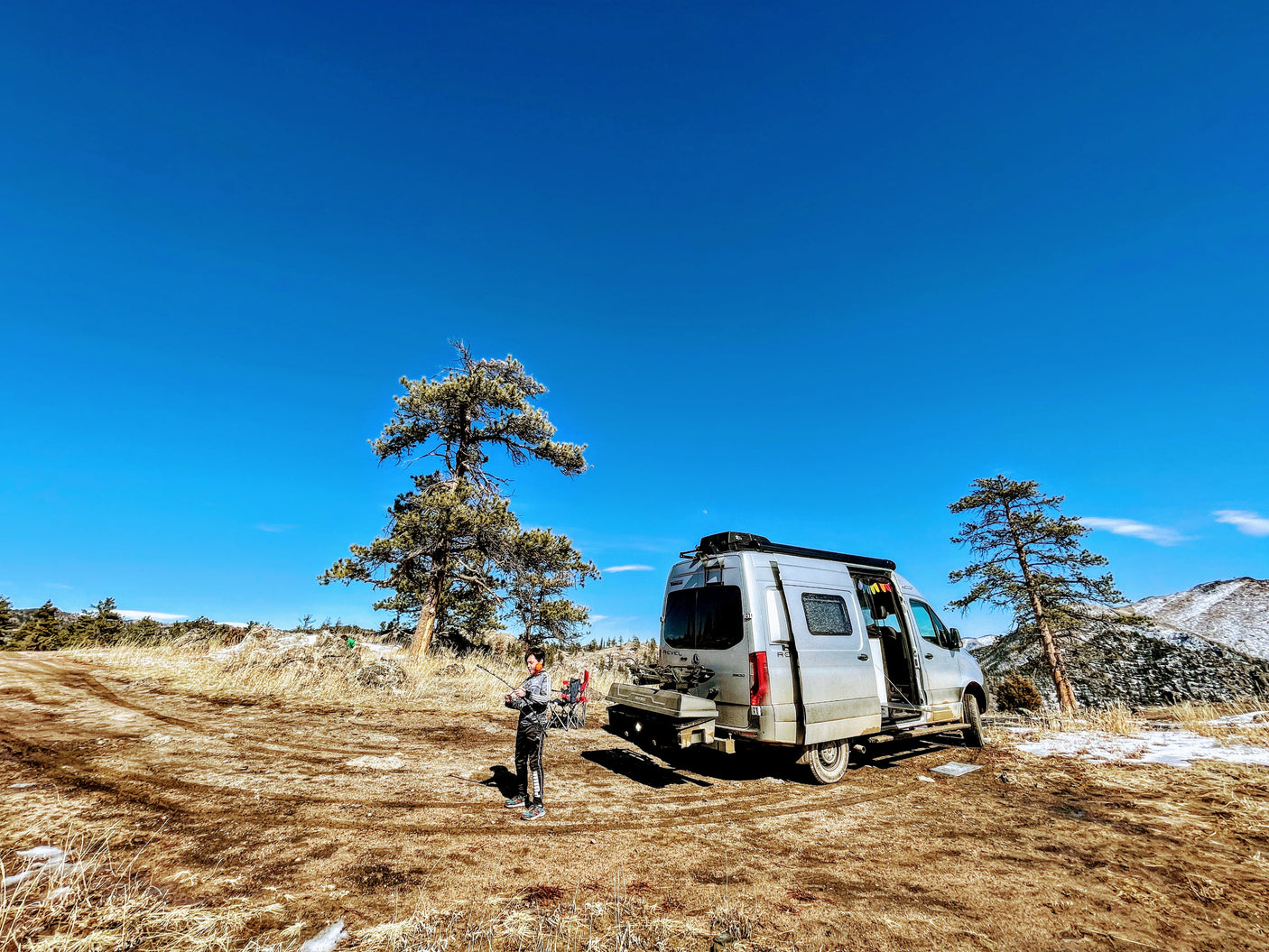 A person stands by a silver RV rental van in a dry, snowy landscape with pine trees and mountains under a blue sky.