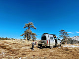 A person stands by a silver RV rental van in a dry, snowy landscape with pine trees and mountains under a blue sky.