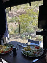Enjoying a meal in an RV rental: Plates of food (omelets, asparagus, salad) on a table, with a view of a rocky desert and a brown sign outside.
