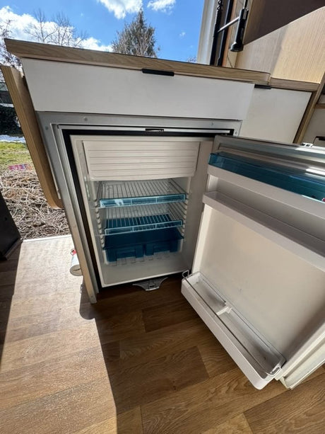 Inside an RV rental: Open refrigerator/freezer with shelves, ice box, & crisper drawers. Wood floor, outdoor view.