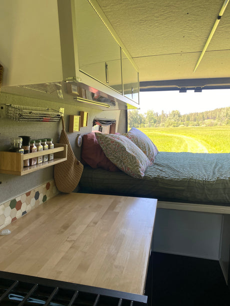 Cozy RV rental interior: kitchen counter, spice rack, bed with nature view, and overhead cabinets for storage.