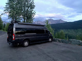 Scenic RV rental: Dark grey camper van parked on a mountain road overlooking forests & peaks under cloudy skies.