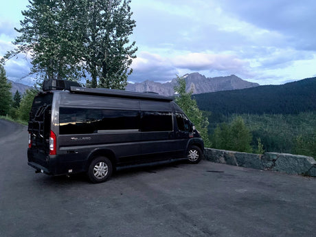 Scenic RV rental: Dark grey camper van parked on a mountain road overlooking forests & peaks under cloudy skies.