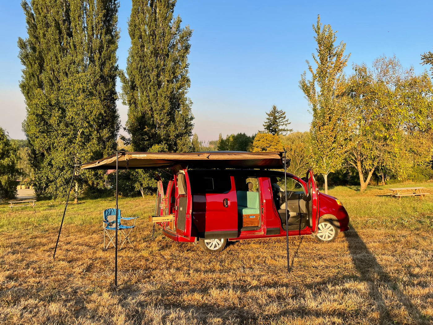 Red camper van RV rental set up with an awning and chair in a sunny field, ready for outdoor adventure. — photo 2