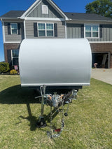 A white, rounded top RV rental trailer with its hitch, parked on a sunny lawn in front of a modern house.