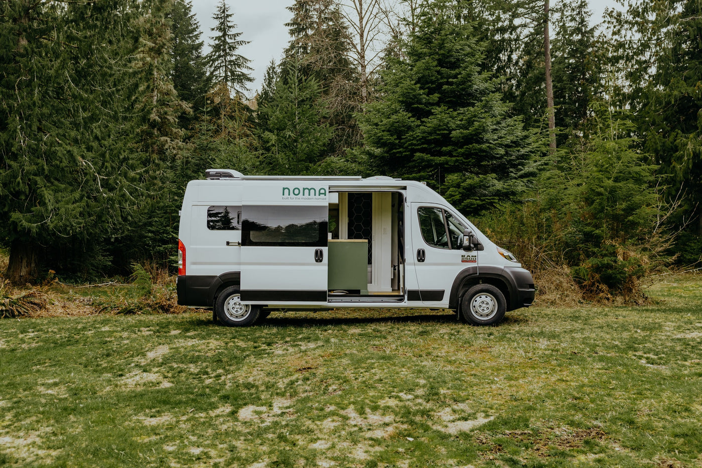 White Noma camper van, an ideal rv rental, parked on grass with its side door open, revealing interior, backed by evergreen trees. — photo 17