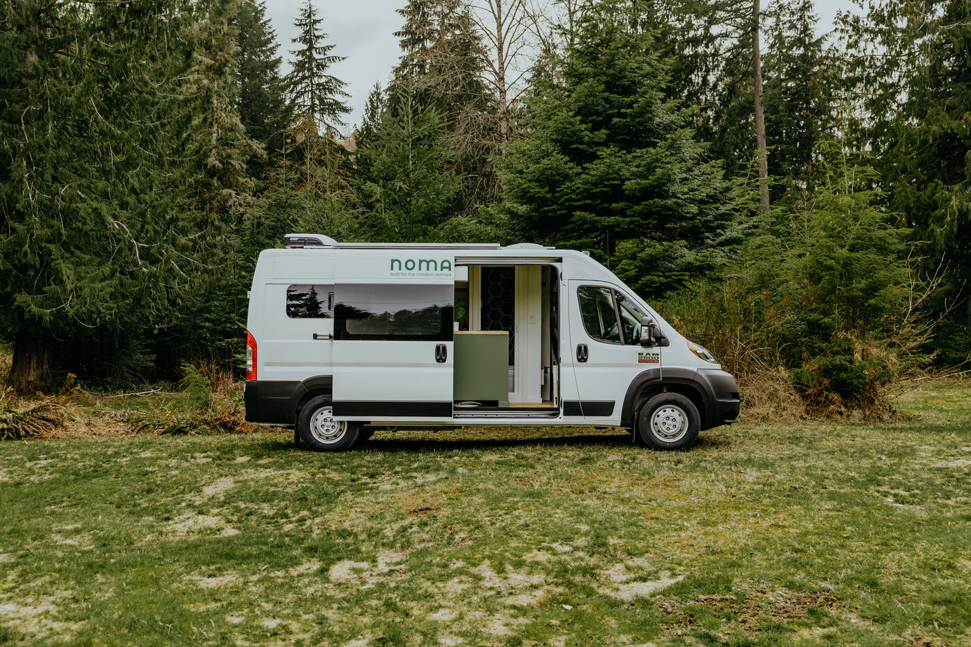 White Noma camper van, an ideal rv rental, parked on grass with its side door open, revealing interior, backed by evergreen trees.