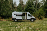 White Noma camper van, an ideal rv rental, parked on grass with its side door open, revealing interior, backed by evergreen trees.