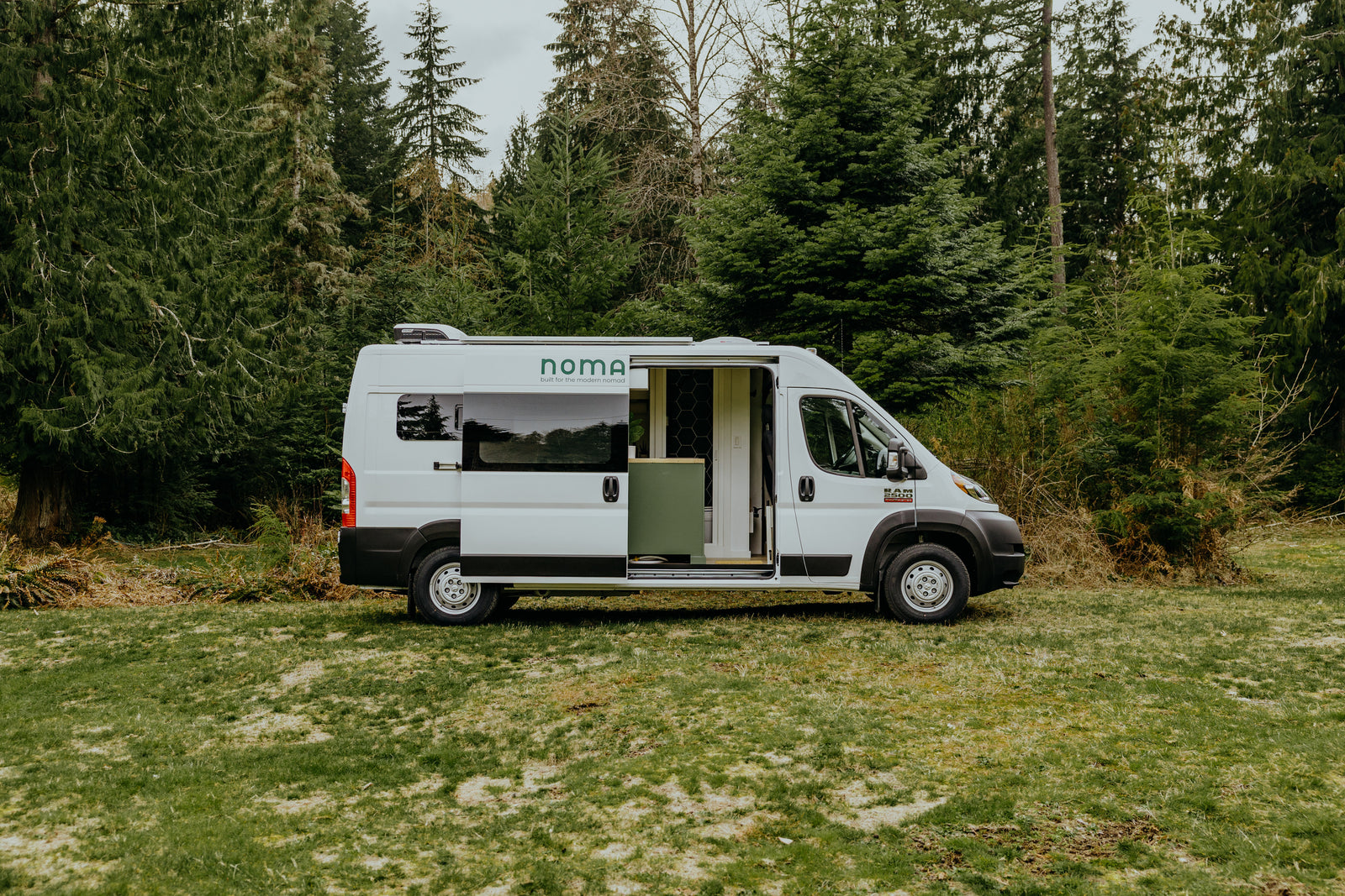 White Noma camper van, an ideal rv rental, parked on grass with its side door open, revealing interior, backed by evergreen trees.