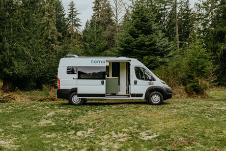 White Noma camper van, an ideal rv rental, parked on grass with its side door open, revealing interior, backed by evergreen trees.