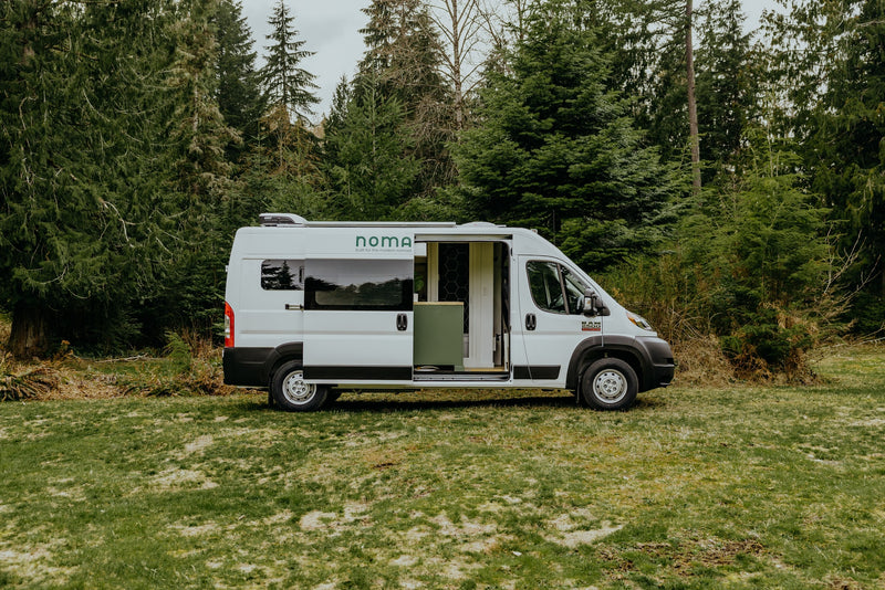 White Noma camper van, an ideal rv rental, parked on grass with its side door open, revealing interior, backed by evergreen trees. — photo 17