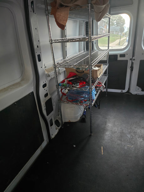 Interior of a white utility van, possibly for rv rental. Chrome shelving unit holds supplies; black textured floor.