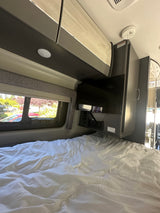 Cozy RV rental interior with a comfortable bed, wall-mounted TV, and window view. Includes overhead storage for travelers.