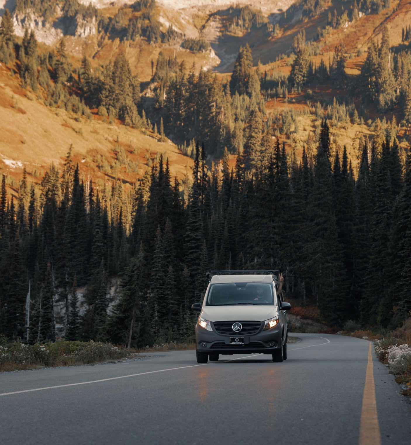 A light Mercedes van, ideal for rv rental adventures, drives a mountain road through dense evergreen forests and sunlit peaks.