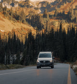 A light Mercedes van, ideal for rv rental adventures, drives a mountain road through dense evergreen forests and sunlit peaks.