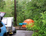 Campsite scene: A blue car and white teardrop RV rental are parked next to an orange tent, surrounded by lush green trees.
