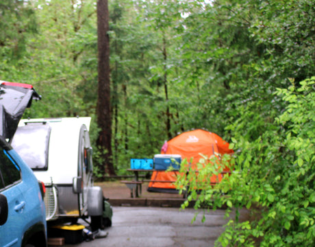 Campsite scene: A blue car and white teardrop RV rental are parked next to an orange tent, surrounded by lush green trees.