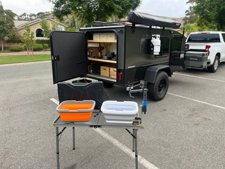 RV rental: Compact overland camper trailer with outdoor kitchen, wash basins, and attached pickup truck, parked in a lot.
