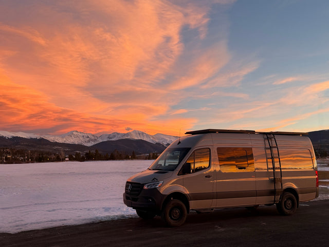 An RV rental Mercedes Sprinter camper van reflecting a vibrant sunset sky, parked against snowy mountains.