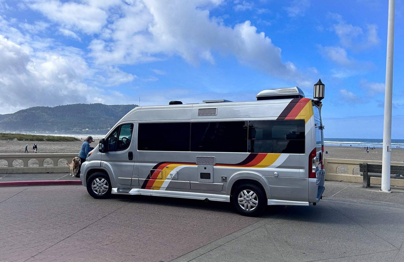 A silver **rv rental** with colorful stripes is parked by a scenic beach, offering travel adventures. A person & dog are near.