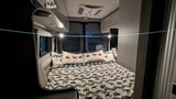 Inside a cozy RV rental bedroom. Features a bed with deer/tree bedding, overhead cabinets, and windows.
