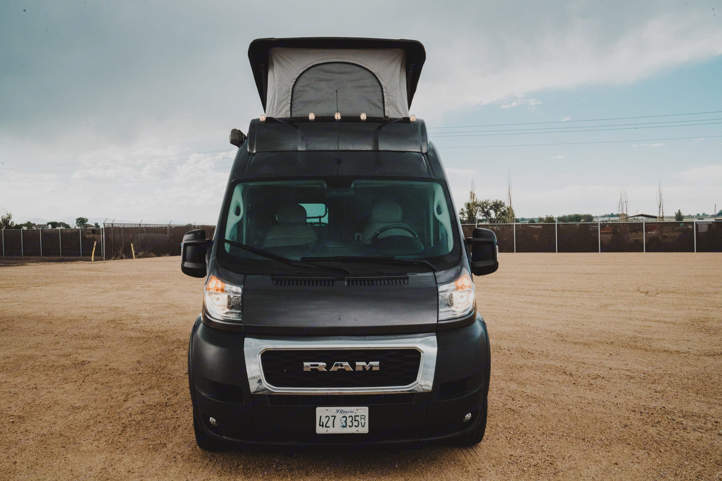 A black Ram ProMaster camper van with a raised pop-top roof, parked on a dirt lot. Explore with this RV rental!