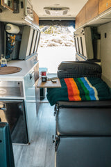An RV rental camper van interior: kitchenette, seating with a rainbow blanket, and a beautiful outdoor view.