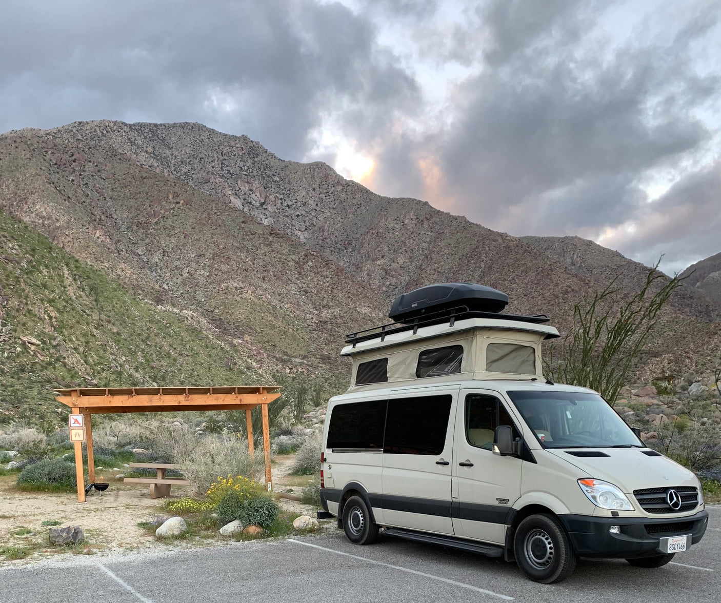 Beige RV rental camper van parked at a desert mountain campsite with a pop-top roof and cargo box, picnic area nearby.