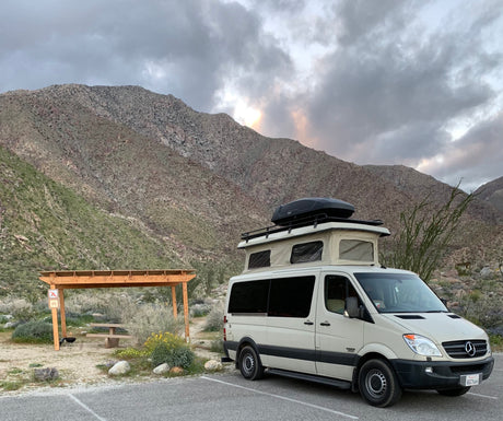 Beige RV rental camper van parked at a desert mountain campsite with a pop-top roof and cargo box, picnic area nearby.