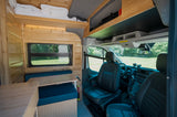 Interior of a cozy RV rental camper van with wooden walls, a blue dinette, overhead storage, and access to the front cabin.