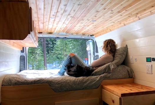 Relaxing RV rental scene. Woman enjoys tranquil snowy forest views from a cozy bed inside a modern camper van.