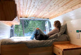 Relaxing RV rental scene. Woman enjoys tranquil snowy forest views from a cozy bed inside a modern camper van.