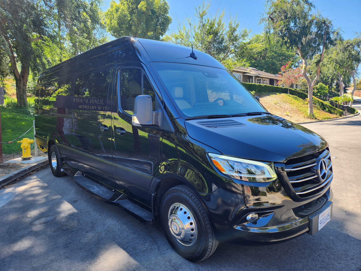 A sleek black Mercedes-Benz Sprinter van, perfect for luxury travel or an RV rental, parked outdoors amidst green trees on a sunny day.