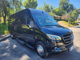 A sleek black Mercedes-Benz Sprinter van, perfect for luxury travel or an RV rental, parked outdoors amidst green trees on a sunny day.