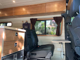 This **rv rental** interior features light wood cabinetry, seating, a fold-out table, and a large window with brown curtains.