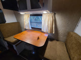 RV rental dinette featuring a wooden table, cushioned benches, window with curtains, and overhead cabinets.