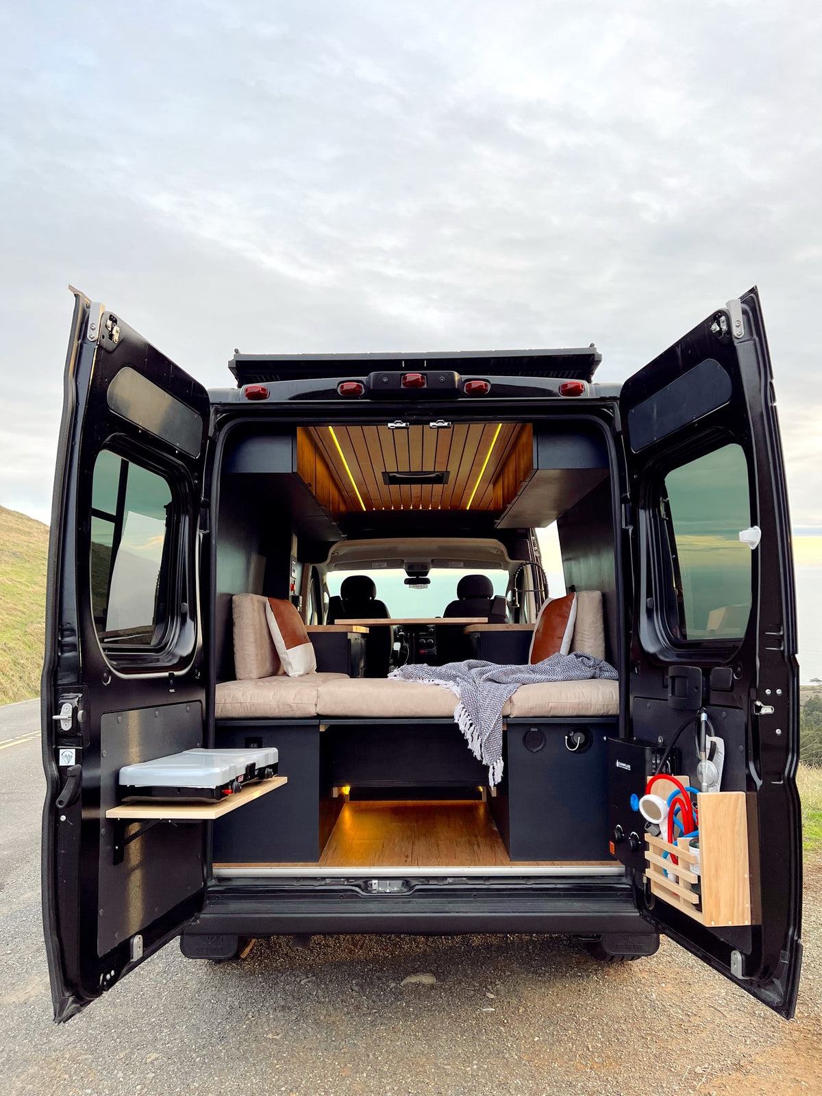 Black RV rental camper van with open rear doors, revealing a custom interior: bed, wooden ceiling lights, kitchen, and outdoor shower. — photo 4
