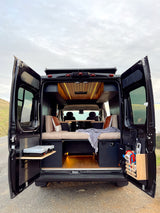 Black RV rental camper van with open rear doors, revealing a custom interior: bed, wooden ceiling lights, kitchen, and outdoor shower.