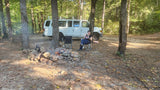 A person relaxes by a campfire at a wooded campsite, beside a white RV rental van. An idyllic outdoor retreat.