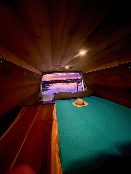 Cozy RV rental interior with a teal bed, wooden walls, and string lights, offering a stunning sunset view over a marina.