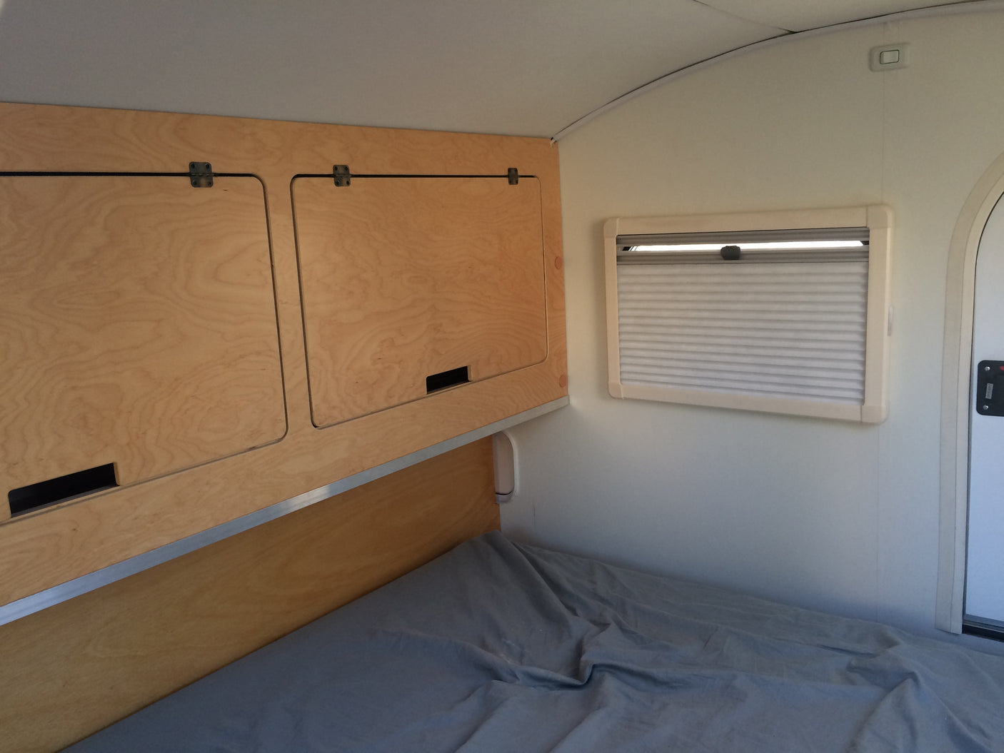 Cozy RV rental interior featuring a bed with gray sheets, wooden overhead cabinets, and a window with blinds.