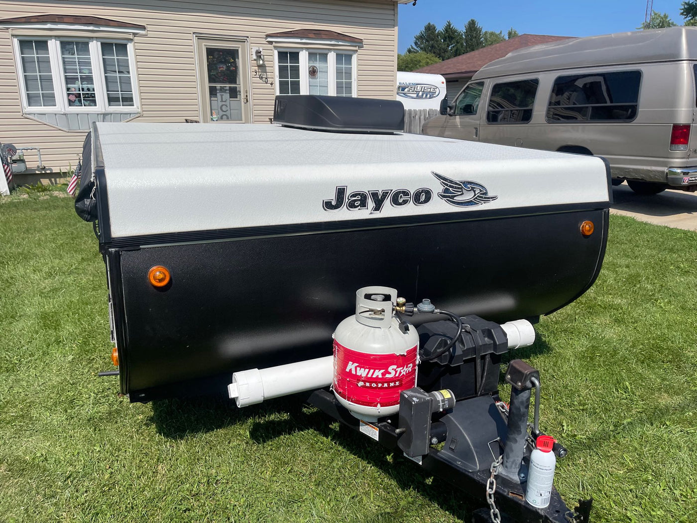 Jayco pop-up RV rental trailer, black and white with propane tank, parked on grass. A house and van are behind. — photo 7