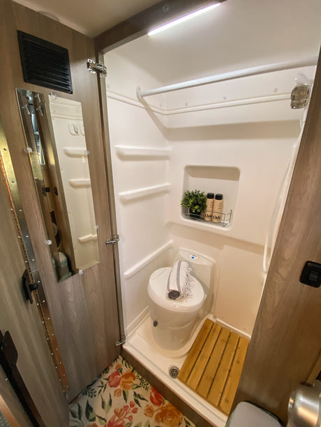 This RV rental wet bath features a compact design with a toilet, shower, shelves for toiletries, and a floral rug.