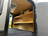 Teardrop RV rental interior: cozy wooden walls, built-in shelves with controls, a comfy bed, and an open door.