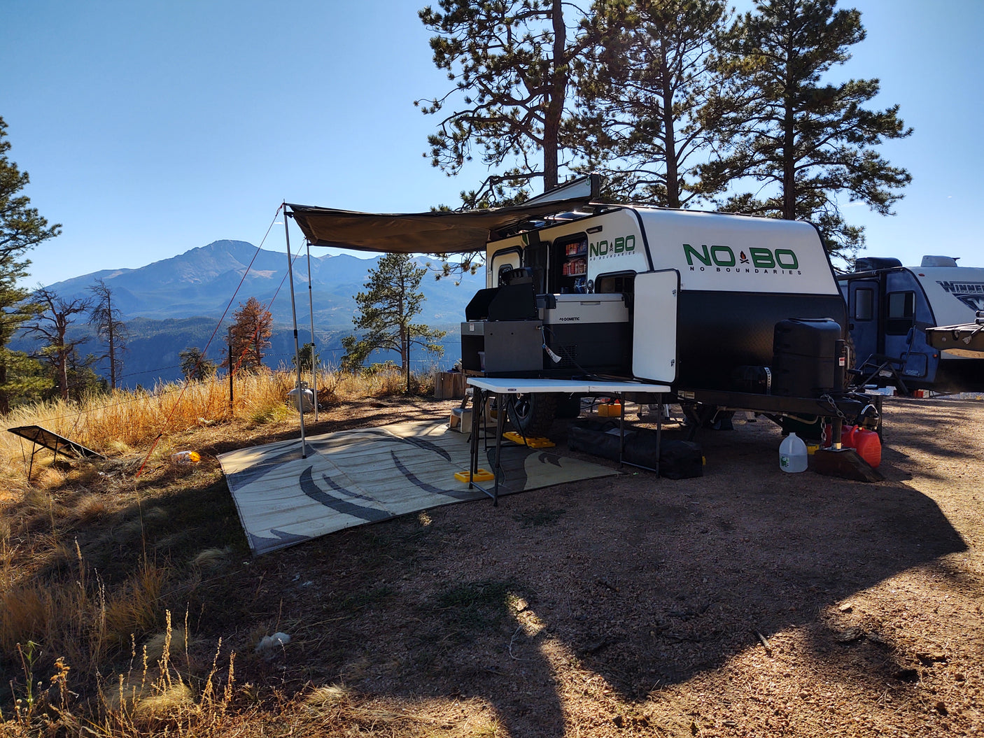 Scenic rv rental camping with a "NO BO" camper, awning, and mountain views. Outdoor kitchen and solar panel visible.