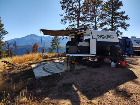 Scenic rv rental camping with a "NO BO" camper, awning, and mountain views. Outdoor kitchen and solar panel visible.