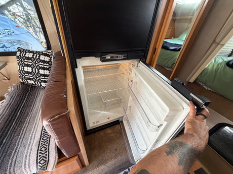 An open, empty refrigerator in an RV rental, showing wire shelves and a person's hand holding the door.