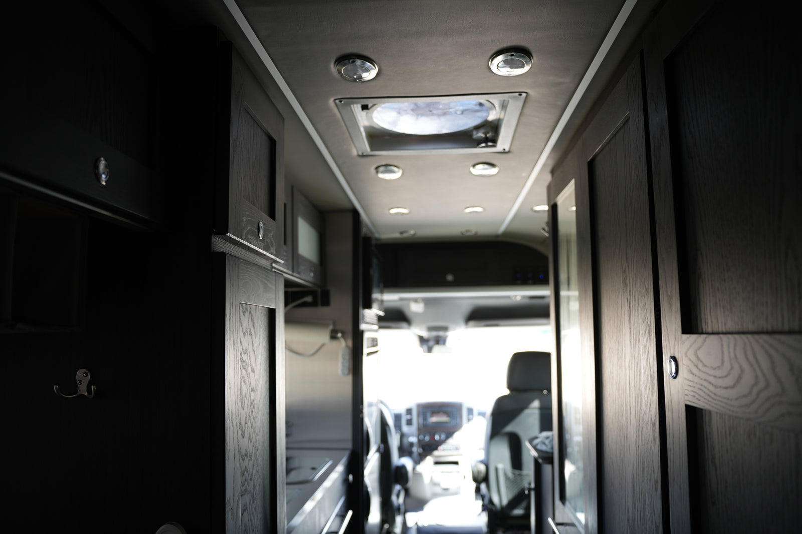 Modern RV rental interior with dark wood cabinets, ceiling lights, and the sunlit driver's area ahead.